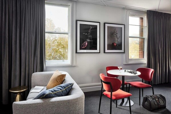 Een moderne hotelkamer met een grijze bank, rode stoelen, een klein tafeltje en twee kunstwerken in lijsten aan de muur.