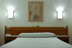 Un dormitorio con cabecera de madera, una cama con ropa de cama blanca, una mesita de noche con lámpara y un cuadro en la pared.