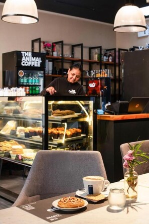 Interior de café com vitrine de produtos de panificação, máquina de café e área de estar com mesa e cadeiras.