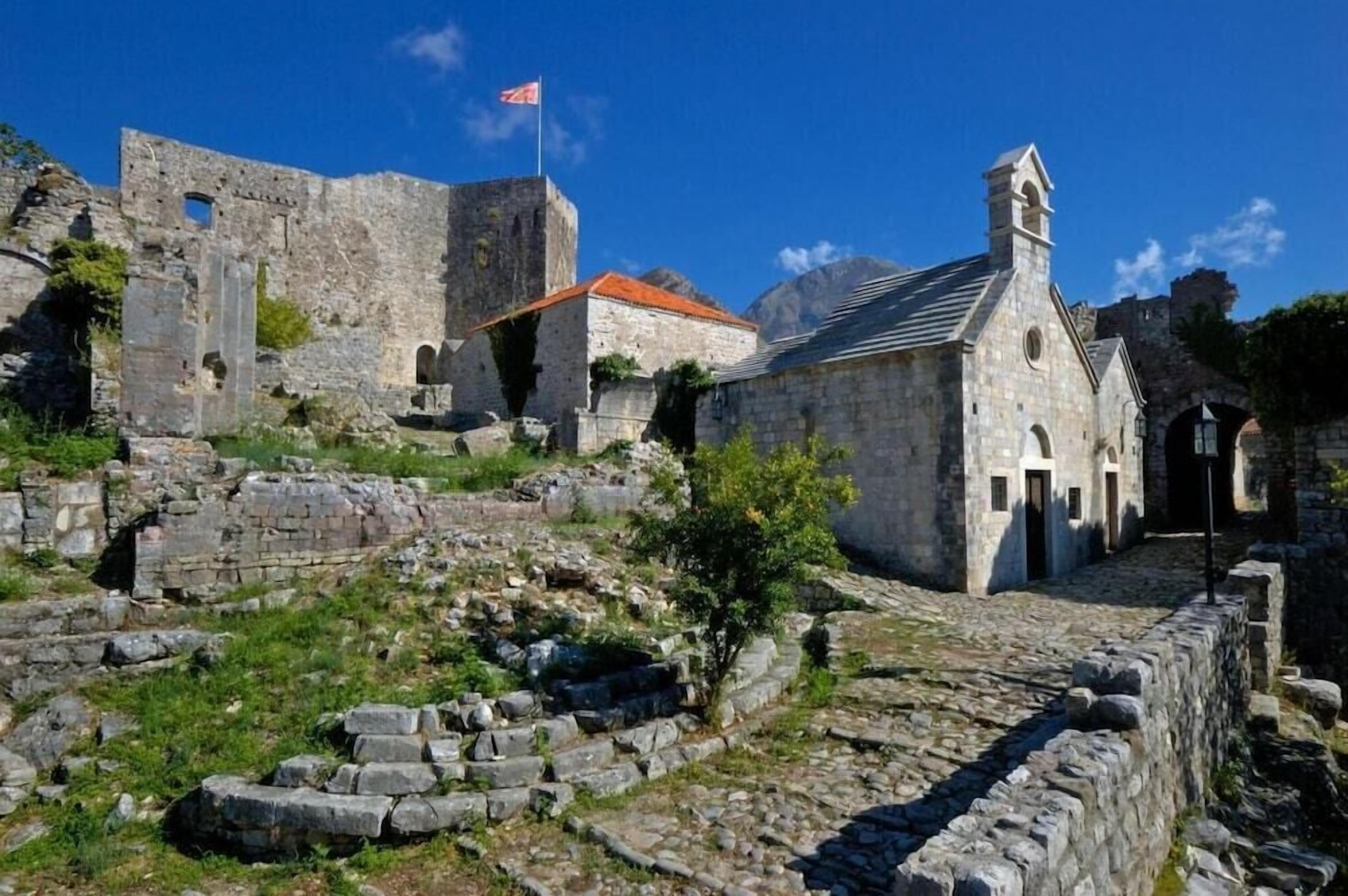 An ancient stone building with a bell tower, surrounded by ruins and a flag.