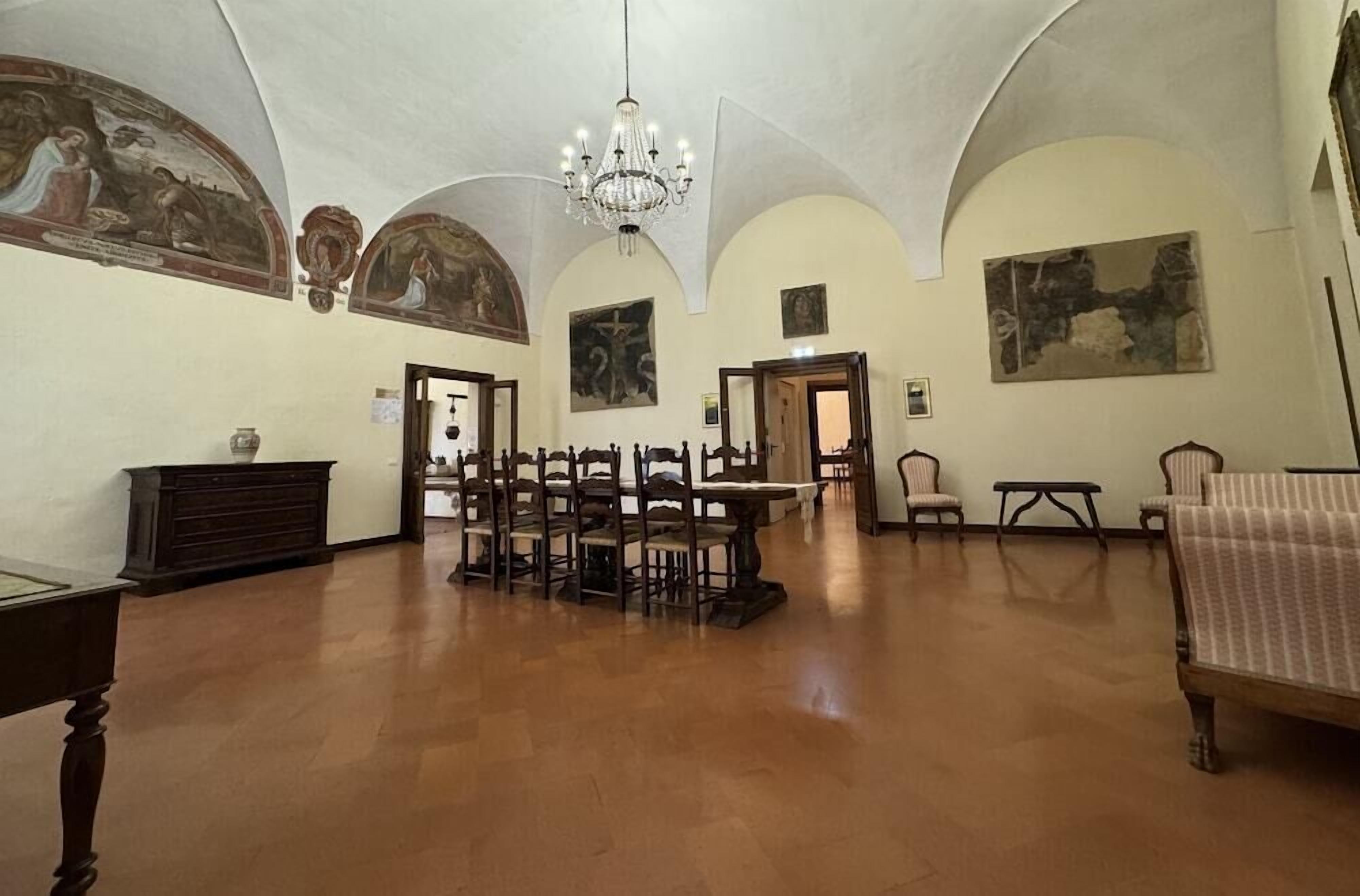Un'ampia stanza con soffitto alto, pavimento in legno e un lampadario. Diverse tavole e sedie in legno sono disposte per la sala da pranzo. Le pareti sono decorate con dipinti classici e affreschi.