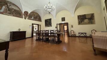 Un'ampia stanza con soffitto alto, pavimento in legno e un lampadario. Diverse tavole e sedie in legno sono disposte per la sala da pranzo. Le pareti sono decorate con dipinti classici e affreschi.
