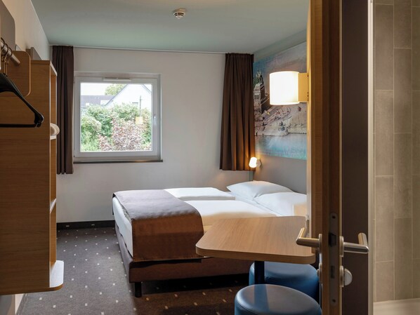 A hotel room with a bed, a small table, a chair, a window with curtains, and a wall-mounted lamp. - B&B Hotel Koblenz (Koblenz)