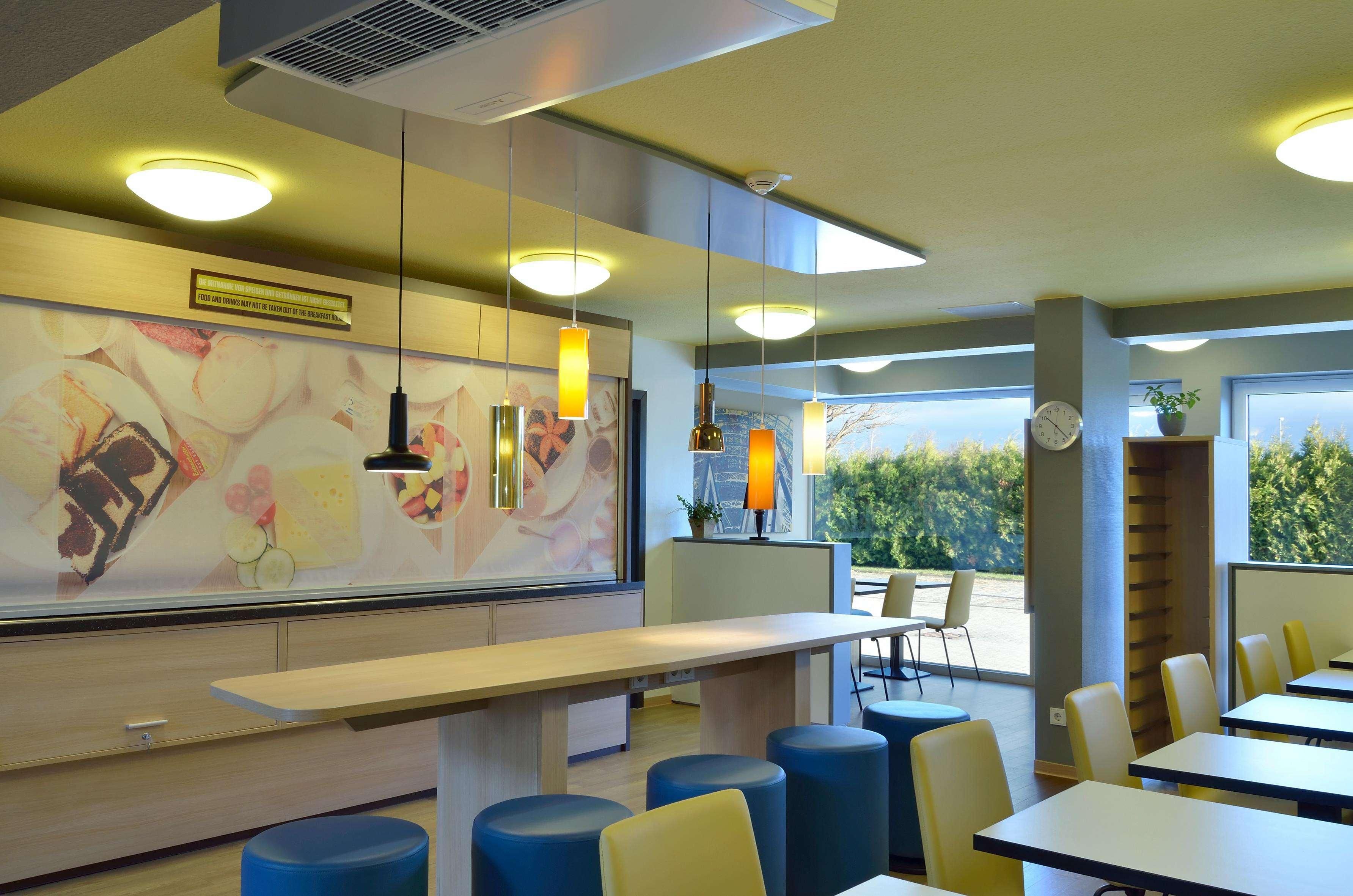 A modern restaurant interior with a long counter, blue and yellow seating, and large windows offering a view of greenery.