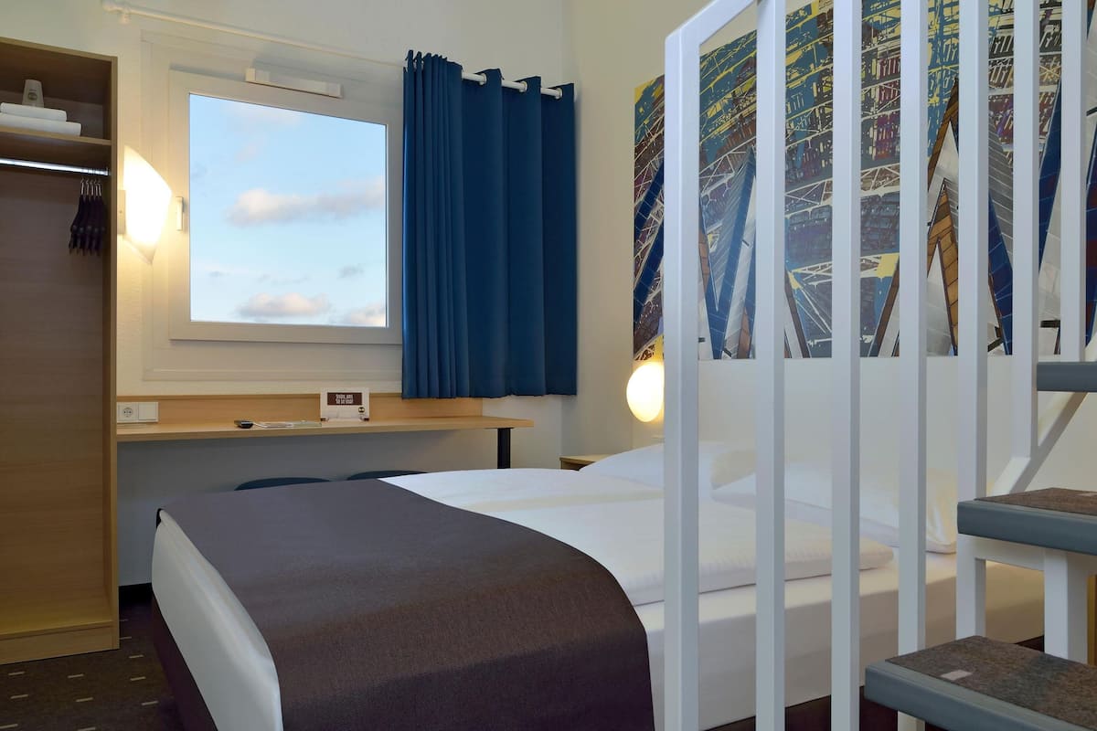 A hotel room with a bed, a desk, a window with curtains, a wall-mounted TV, and a poster.
