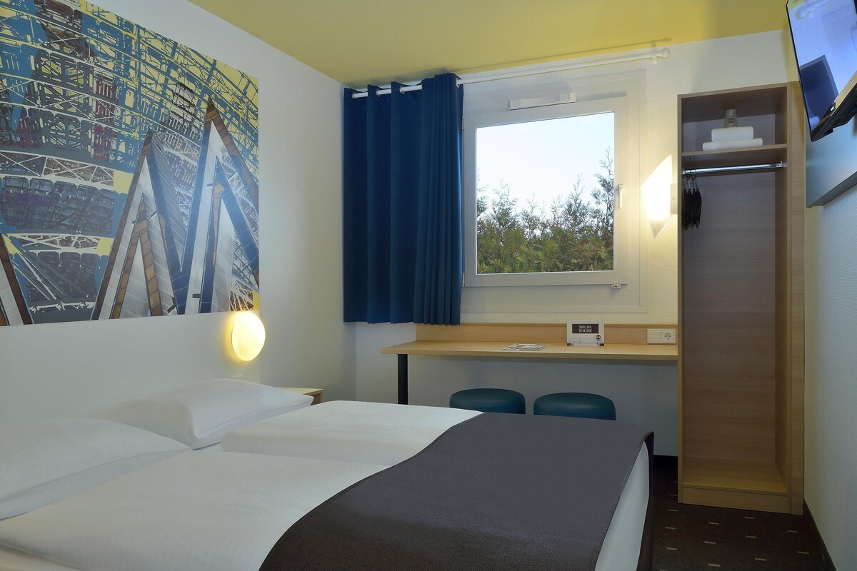 A hotel room with a bed, a desk, two chairs, a window with curtains, a TV, and a painting on the wall.