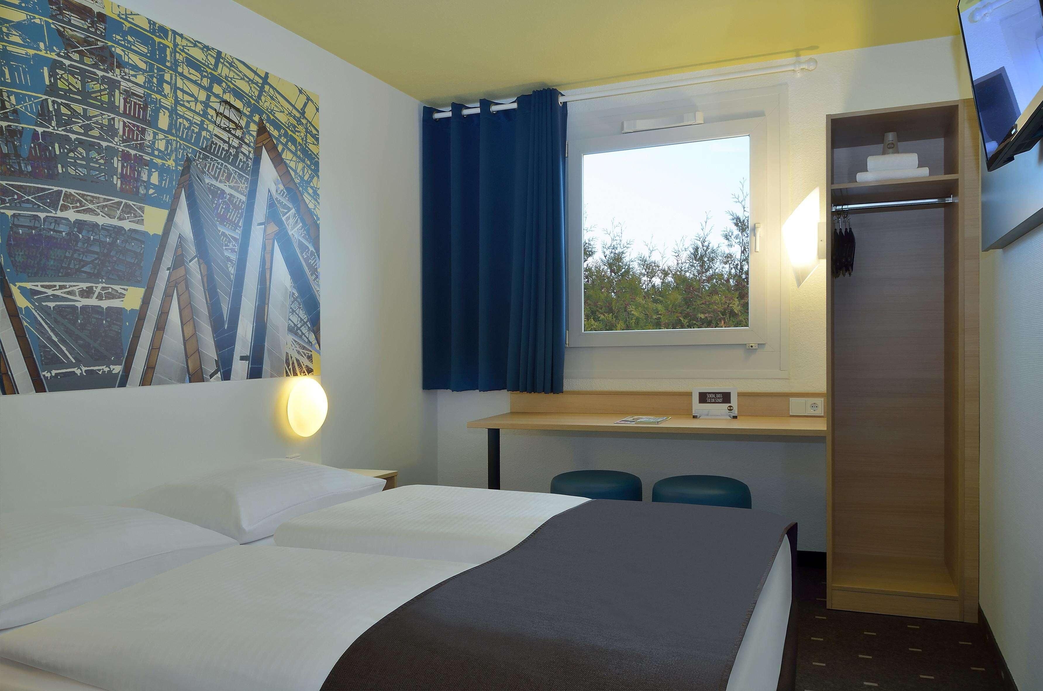 Quarto de hotel com uma cama, uma escrivaninha com duas cadeiras, uma janela com cortinas, uma TV na parede e um quadro na parede.