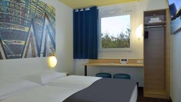 Quarto de hotel com uma cama, uma escrivaninha com duas cadeiras, uma janela com cortinas, uma TV na parede e um quadro na parede.