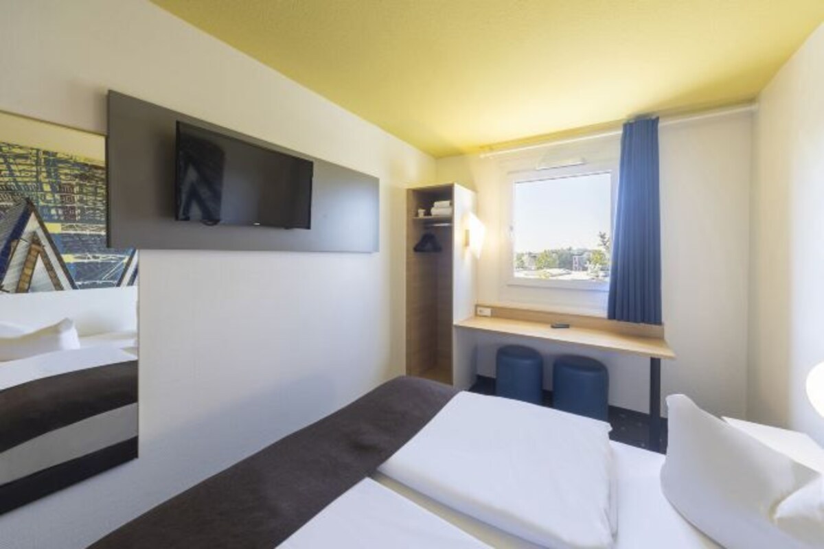 A hotel room with a bed, a desk, a chair, a window with curtains, and a mounted artwork on the wall.