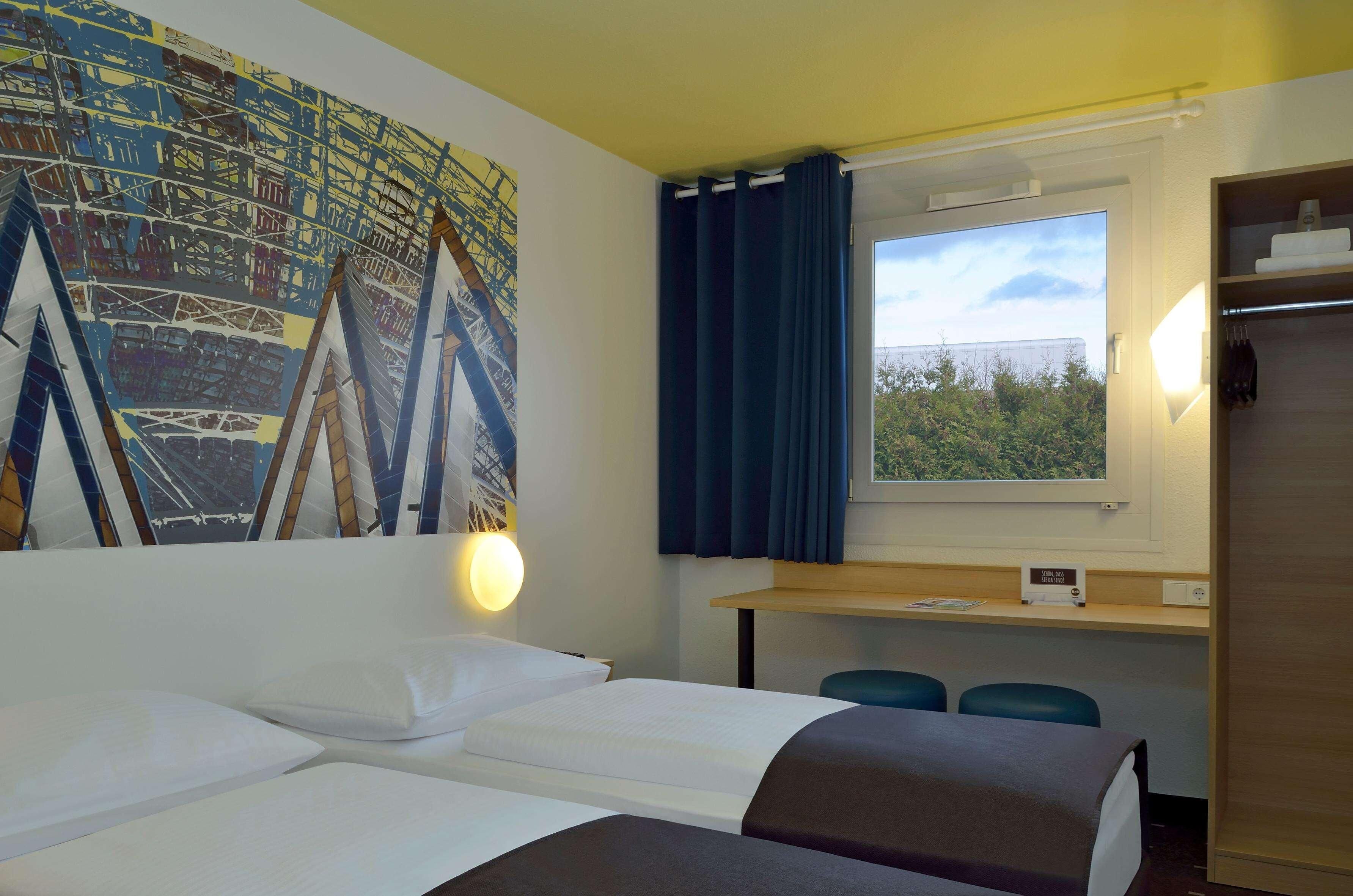 A hotel room with two beds, a desk, a mirror, and a window with a view of greenery.