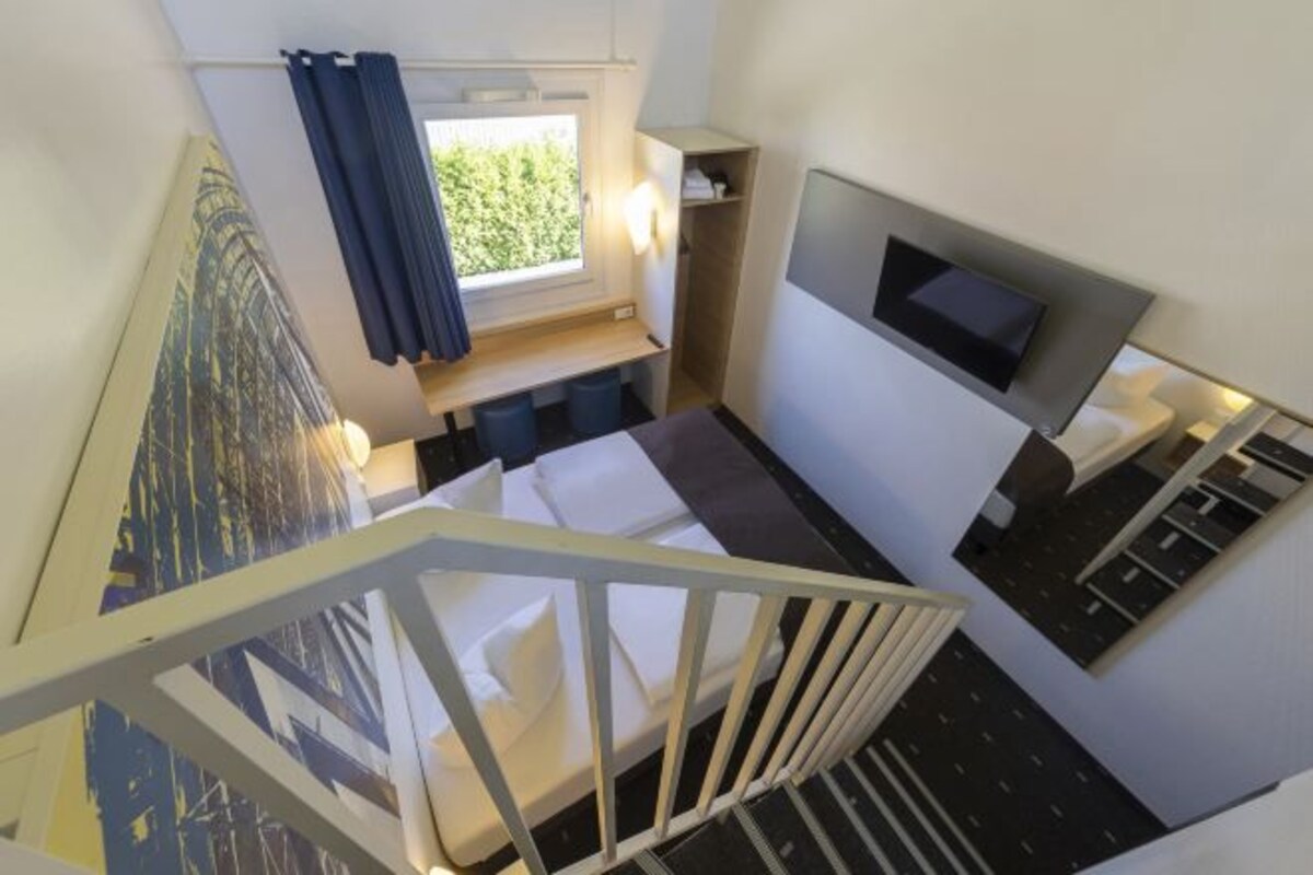 A modern hotel room with a bed, a TV, a window with curtains, and a staircase.