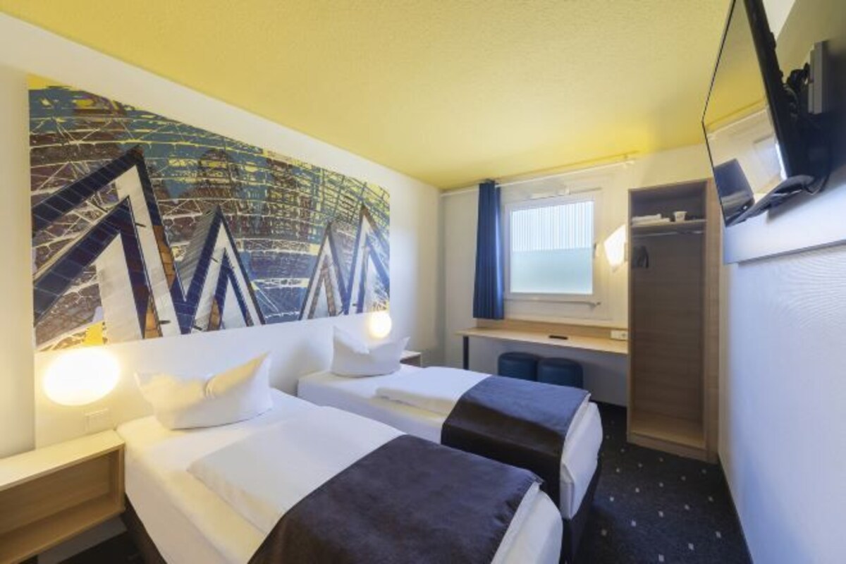 A hotel room with two beds, a desk, a chair, a TV, and a large abstract painting on the wall.