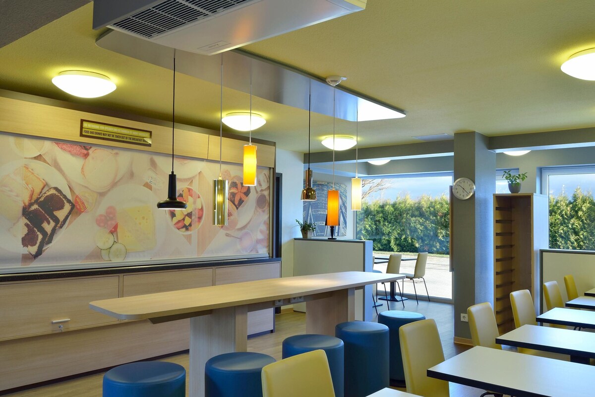 A modern restaurant interior with a long counter, blue and yellow seating, and large windows offering a view of greenery.