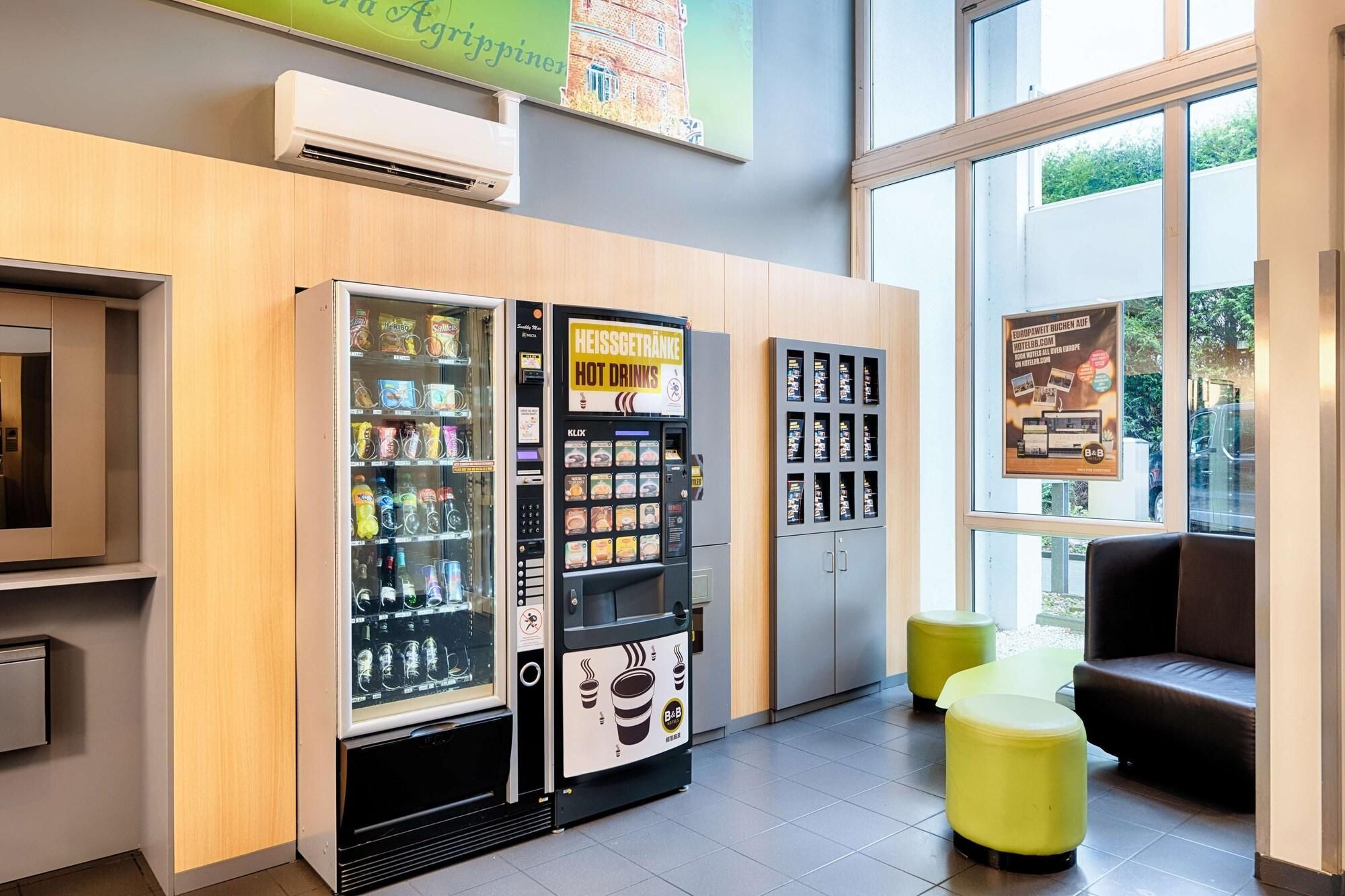 Un hall de hotel con máquinas expendedoras que ofrecen aperitivos y bebidas, una zona de estar con taburetes amarillos y un ventanal con vistas al exterior.