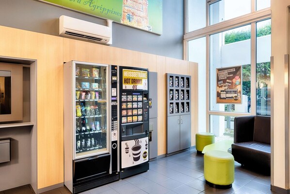 A hotel lobby with vending machines selling snacks and drinks, a seating area with yellow stools, and a large window offering a view of the outdoors. - B&B Hotel Köln-frechen (Frechen)
