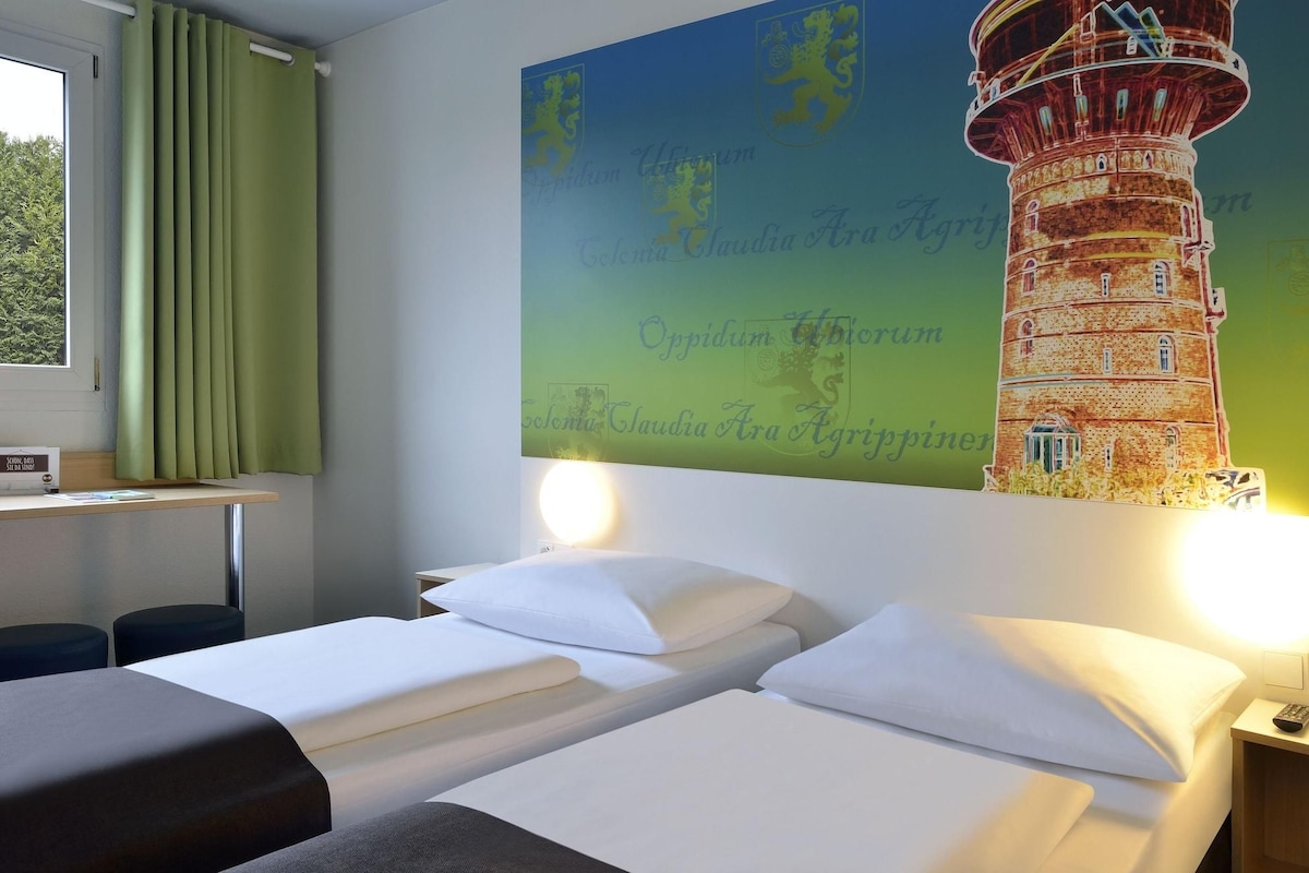 Quarto de hotel com duas camas, uma mesa com abajur, janela com cortinas e um mural de farol na parede.