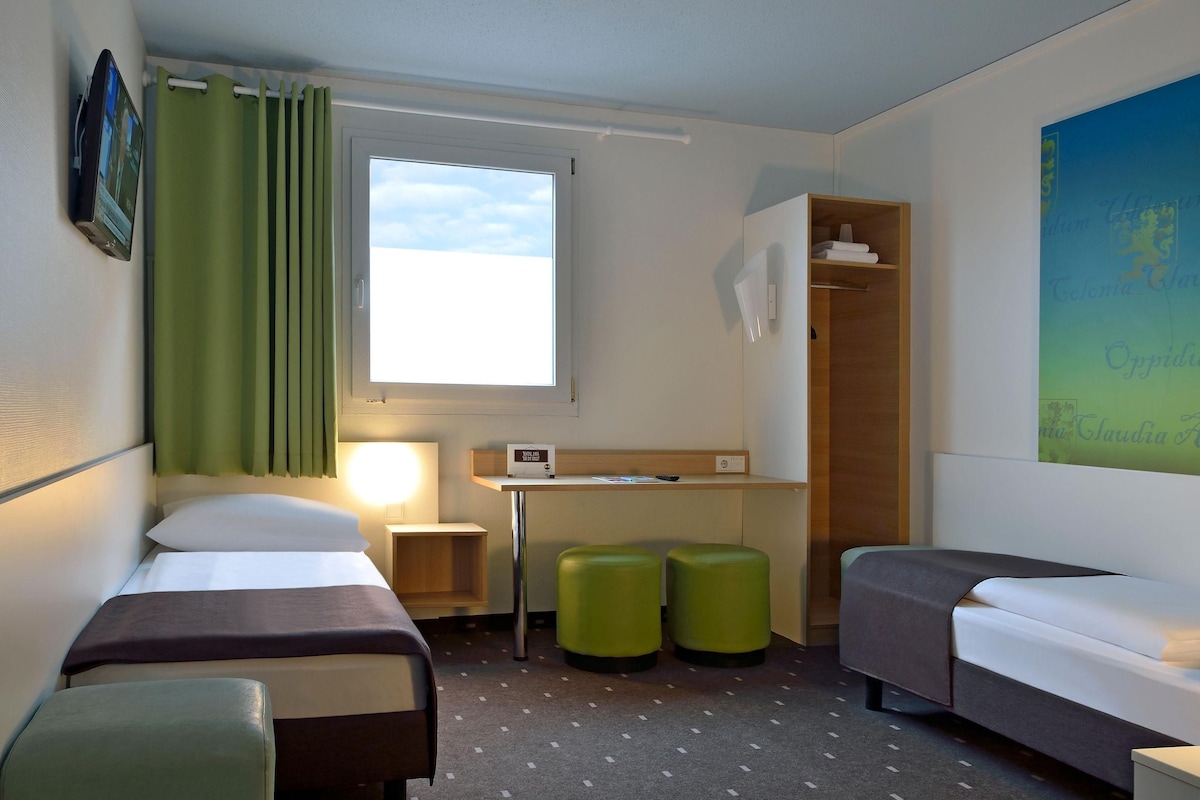 Quarto de hotel com duas camas de solteiro, uma escrivaninha, uma cadeira, dois pufes verdes, um guarda-roupa e uma janela grande com cortinas.