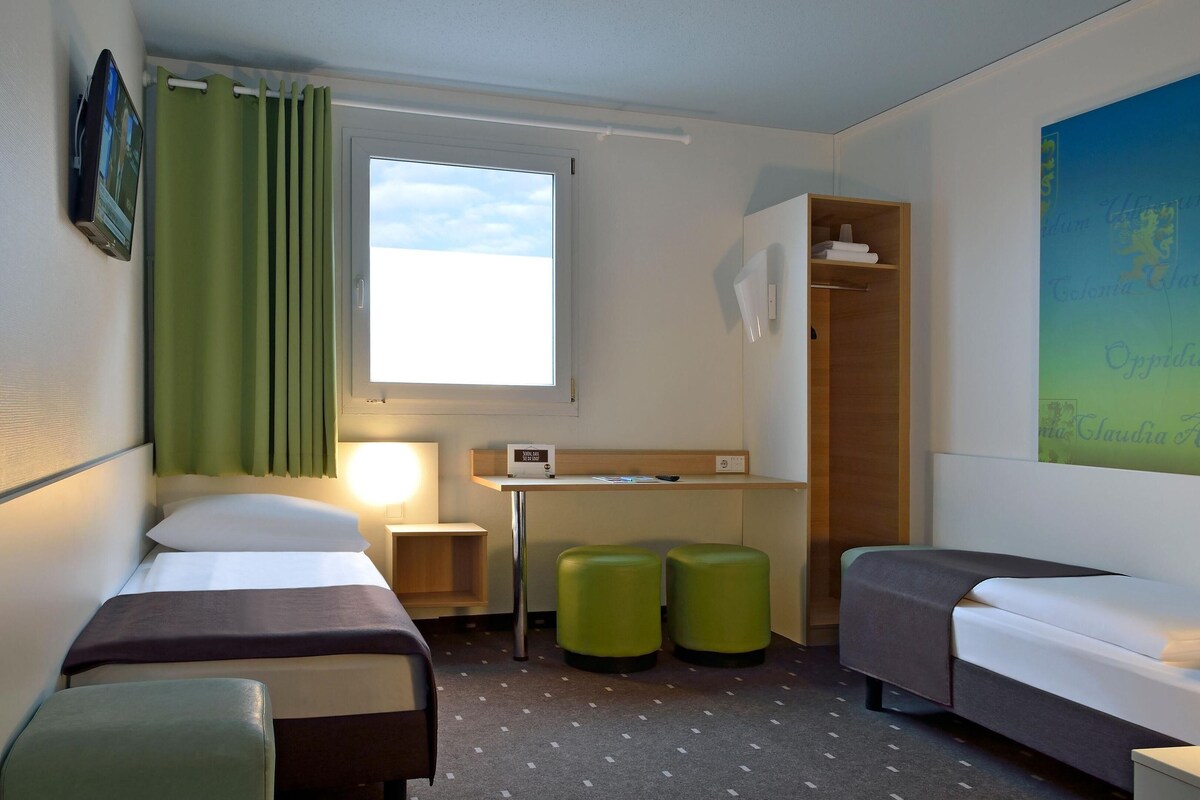 Une chambre d’hôtel avec deux lits simples, un bureau, une chaise, deux poufs verts, une armoire et une grande fenêtre avec des rideaux.