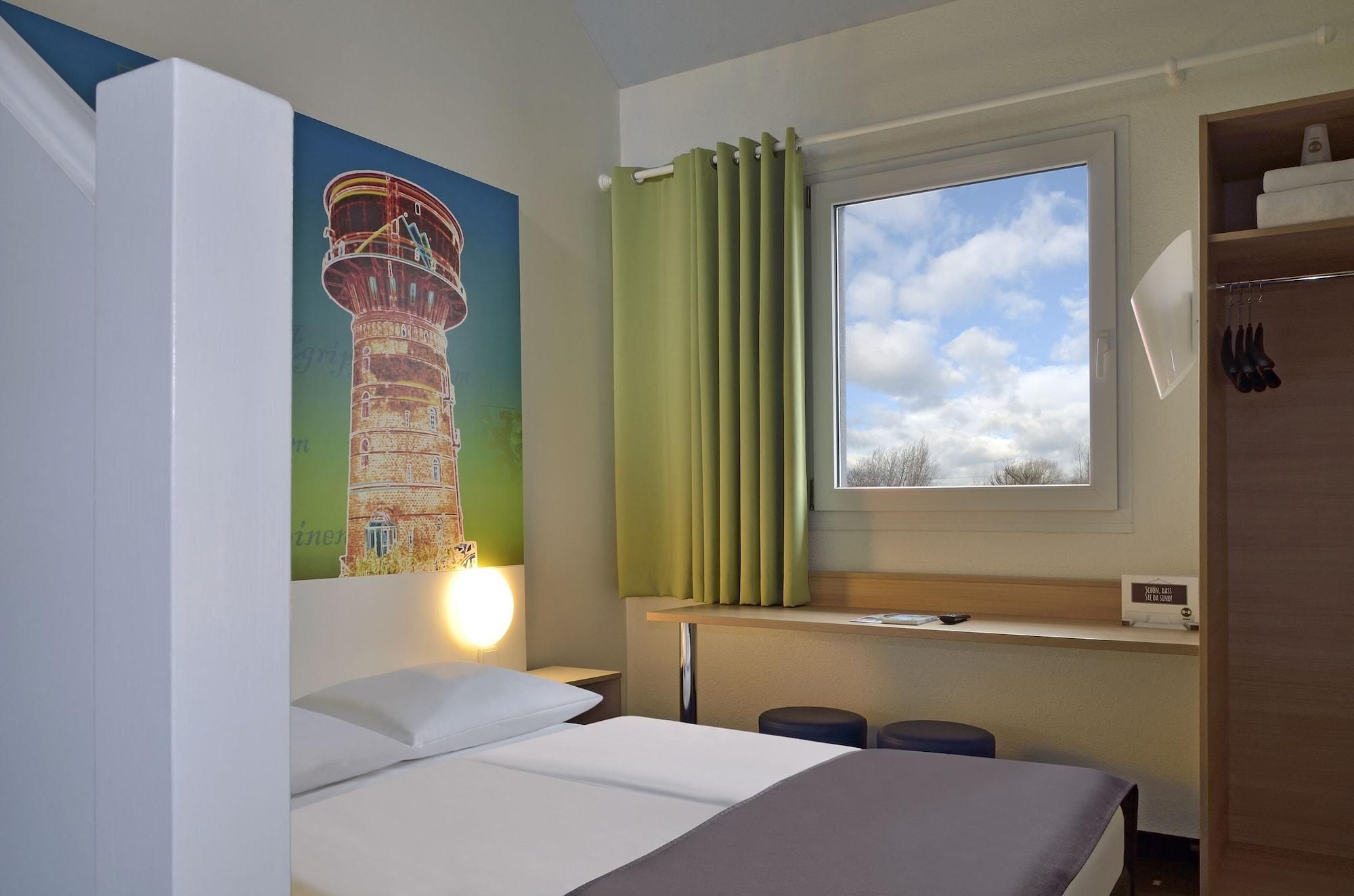 Een hotelkamer met een bed, nachtkastje, raam met gordijnen, een wandlamp en een schilderij van een toren.