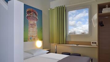Een hotelkamer met een bed, nachtkastje, raam met gordijnen, een wandlamp en een schilderij van een toren.