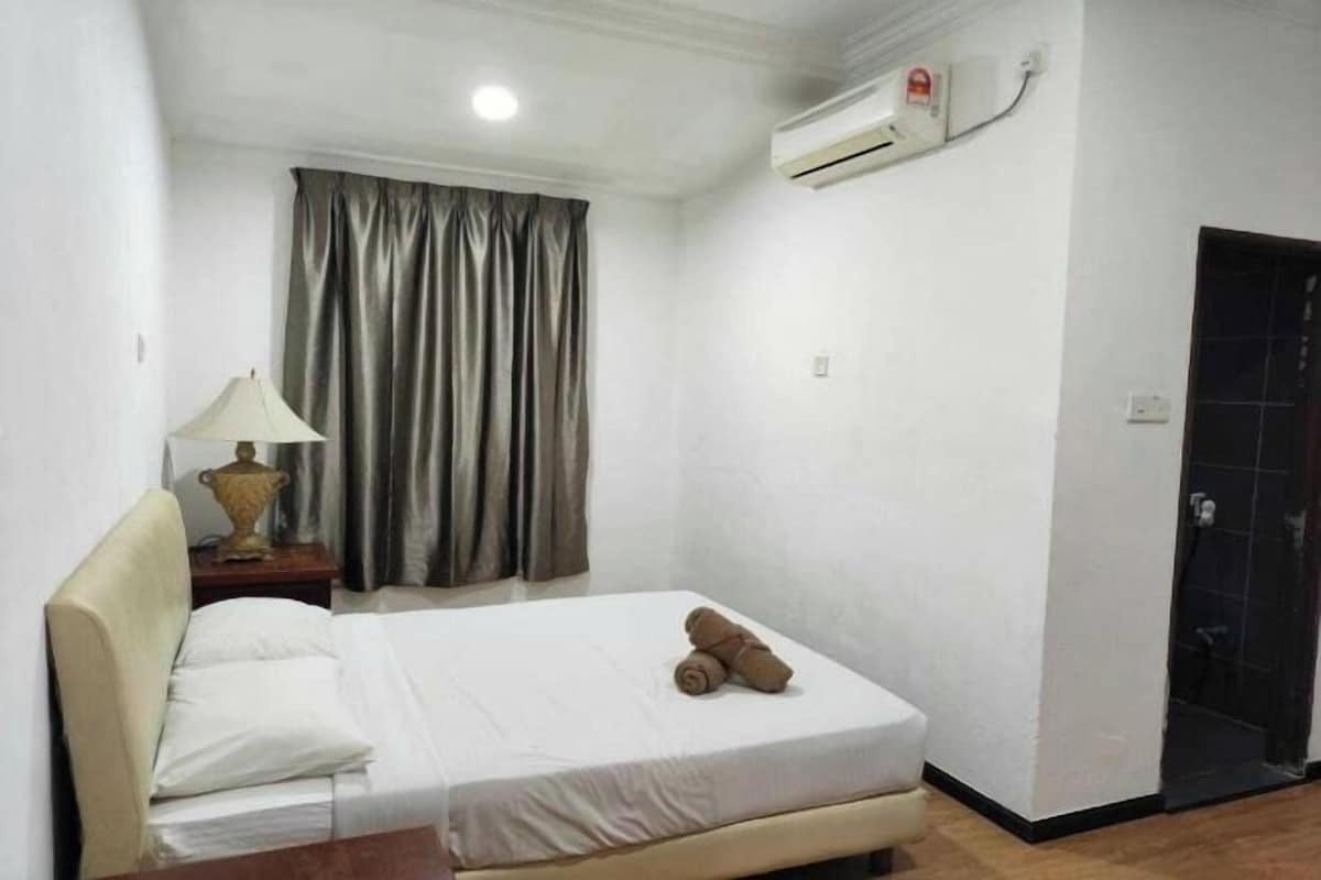 A small, clean room with a bed, a lamp, a window with curtains, and an air conditioning unit.