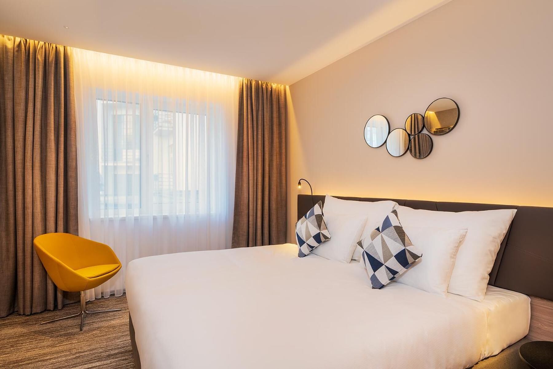 A hotel room with a large bed, a yellow chair, and a view through sheer curtains.