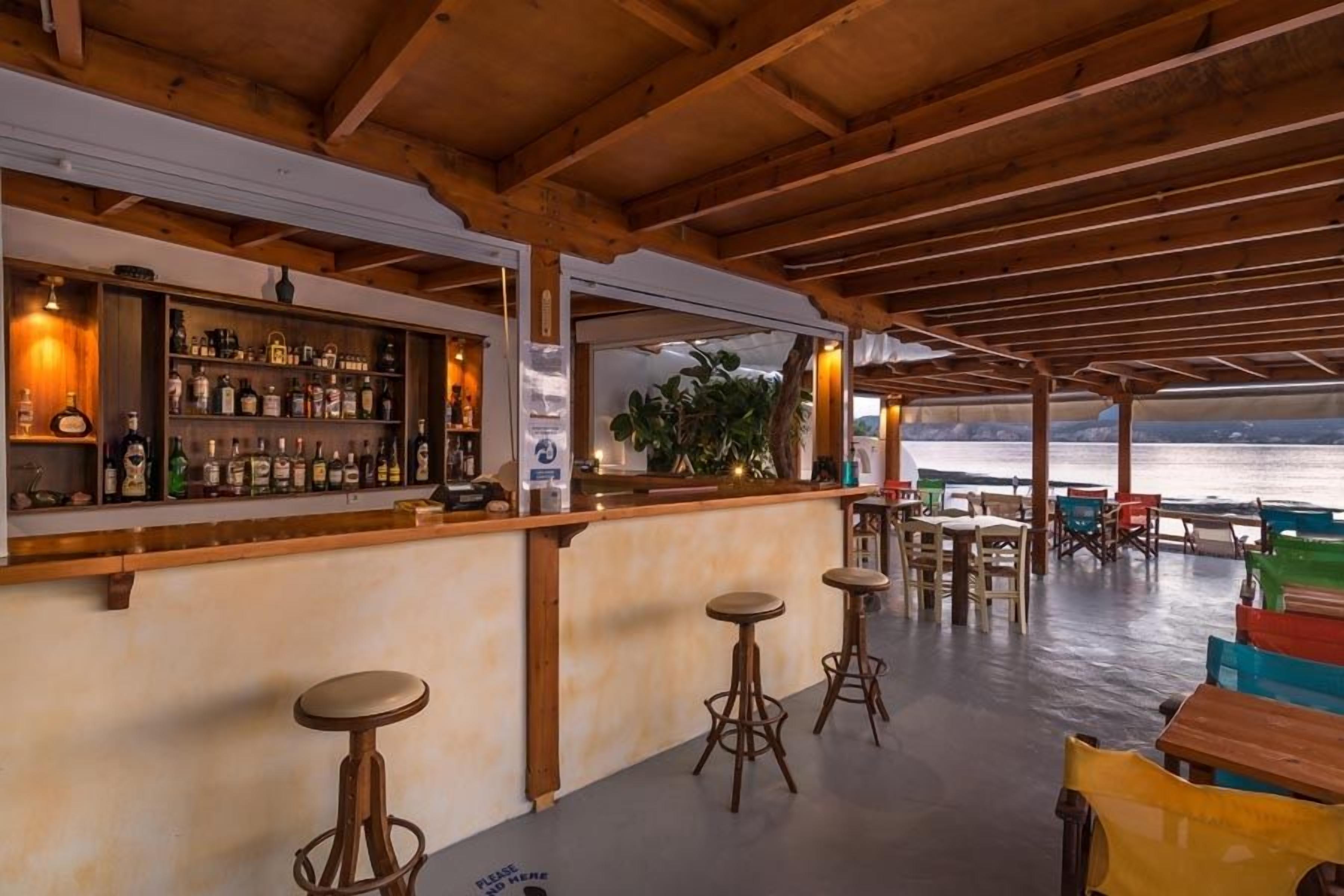 A bar area with wooden beams, a bar counter with bottles, stools, and a view of the sea.