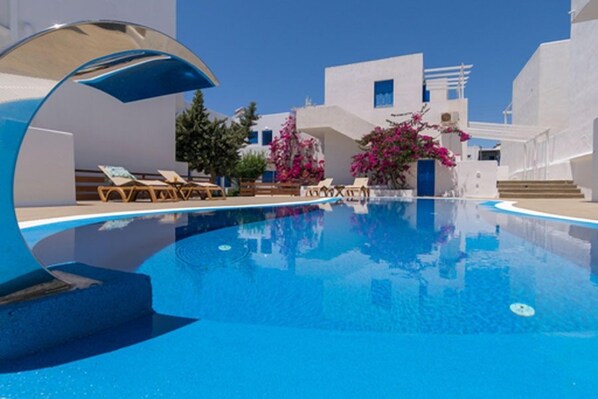 Une piscine d’un bleu éclatant, entourée de bâtiments blancs, avec des chaises longues et des plantes de bougainvilliers aux fleurs roses.