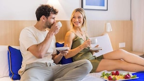 A couple sitting on a bed in a hotel room, holding cups and plates, with a fruit platter on the bed.