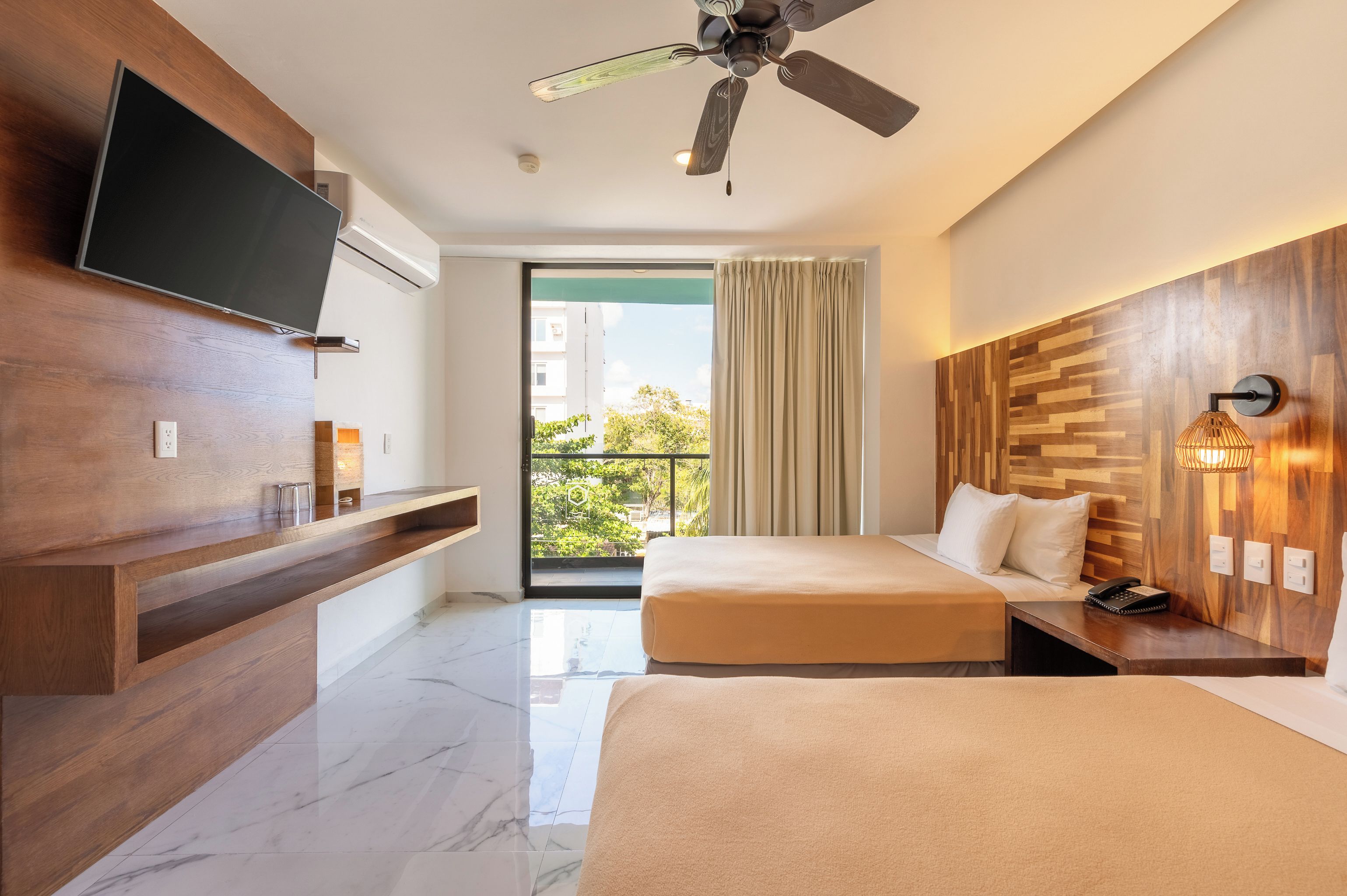 Habitación de hotel con televisión, ventilador de techo, dos camas, balcón con cortinas y diseño de pared de madera.