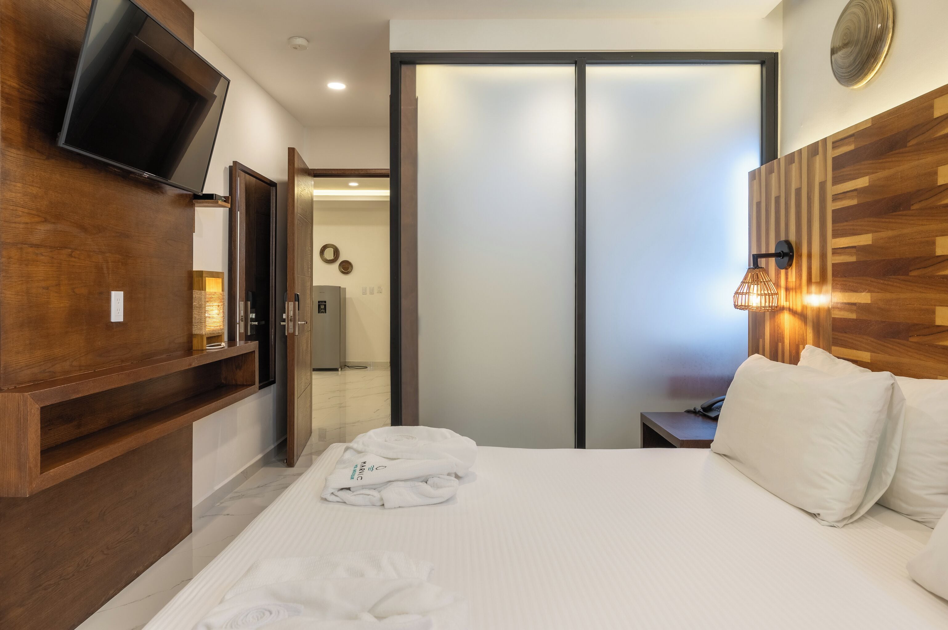 A hotel room with a bed, a TV mounted on the wall, a wooden headboard, a bedside table, and a sliding door closet.