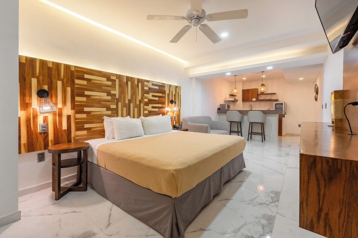 A modern bedroom with a large bed, a wooden accent wall, a ceiling fan, and a kitchenette in the background.