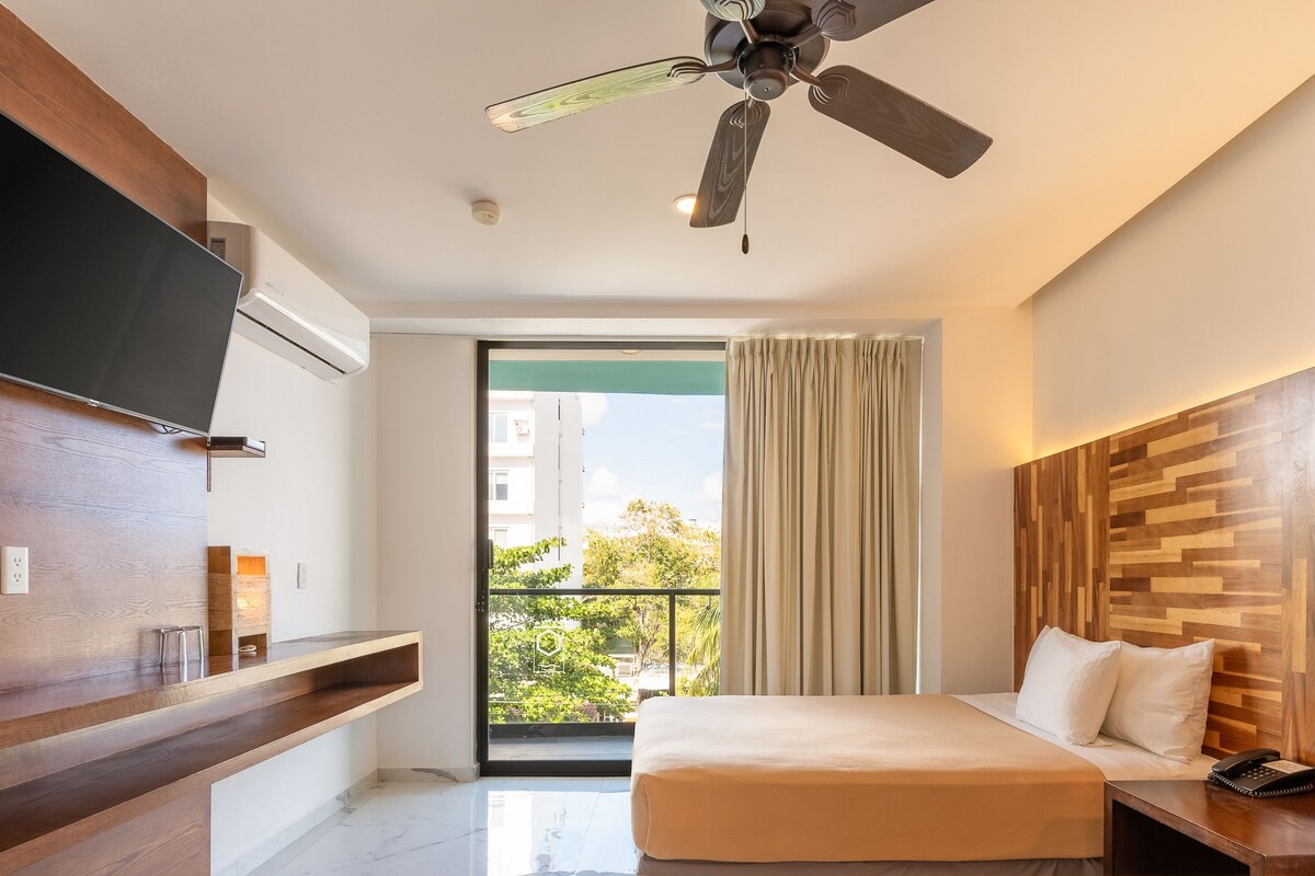 A modern hotel room with a bed, a TV, a ceiling fan, a balcony with curtains, and a wooden accent wall.