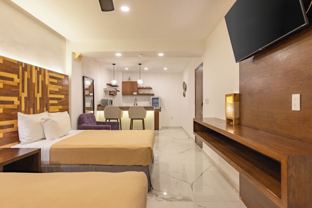 A hotel room with two beds, a TV, a desk with chairs, a kitchen area, and a wooden wall.