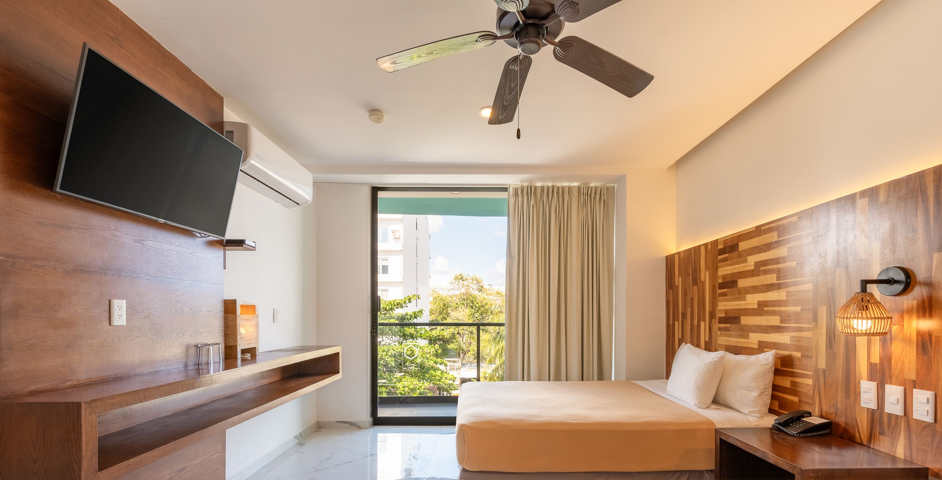 A modern hotel room with a bed, a TV, a ceiling fan, a balcony with curtains, and a wooden accent wall.