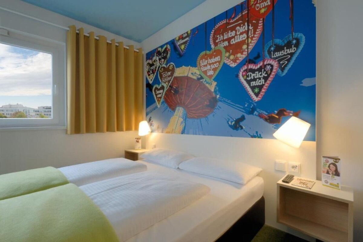 Habitación de hotel con cama, dos mesitas de noche, ventana con cortinas y un mural en la pared con adornos en forma de corazón.