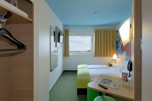 Une chambre d’hôtel comprenant un lit, un bureau, une chaise, un porte-manteau, une fenêtre avec des rideaux et une vue sur la ville.