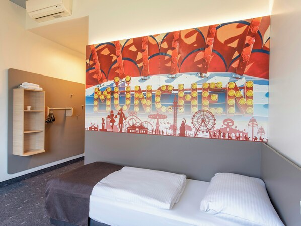 Une chambre d’hôtel avec un lit, une étagère en bois où sont posées des serviettes et un mur orné d’une fresque colorée représentant des monuments emblématiques de Munich.