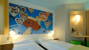 Habitación de hotel con dos camas, un gran mural con motivos de corazón en la pared, lámparas de noche, teléfono en la mesita y un taburete verde.