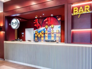 Eine Rezeption mit einem B&B Hotels Schild, einem großen 'BAR'-Schild und einem farbenfrohen Hintergrund mit Zitronen.
