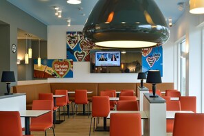 Ein modernes Restaurant mit orangefarbenen Sitzbänken und Tischen, einer großen Pendelleuchte und einem Fernsehbildschirm, der eine Nachrichtensendung zeigt.