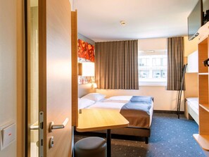Ein kleines Hotelzimmer mit einem Einzelbett, einem kleinen Schreibtisch, einem Stuhl und einem Fenster mit Vorhängen.