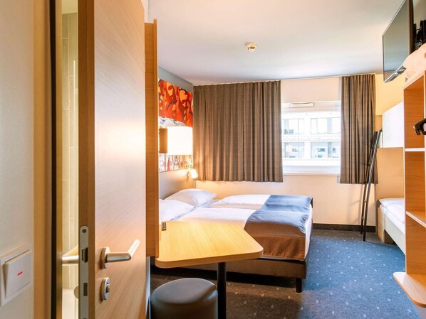 Une chambre d’hôtel de taille modeste, comprenant un lit simple, un petit bureau, une chaise et une fenêtre avec des rideaux.