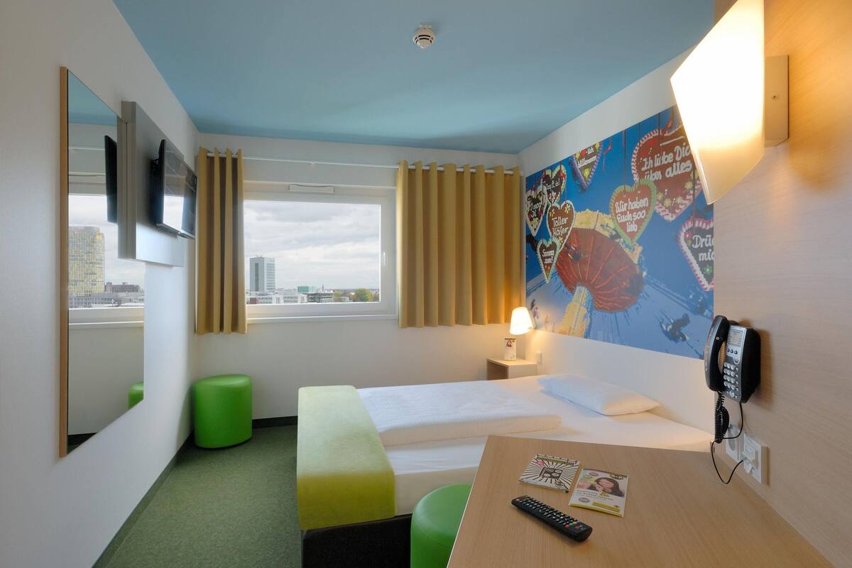 Habitación de hotel con cama, escritorio, reposapiés verde, televisor, lámpara, teléfono y ventana con cortinas.