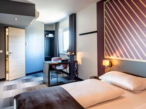 Ein Hotelzimmer mit einem Bett, einem Schreibtisch mit Stuhl, einem kleinen Tisch und einem Fenster mit Vorhängen.