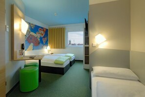 Ein Hotelzimmer mit zwei Betten, einem Schreibtisch, einem grünen Hocker, einem Fenster mit Blick auf die Stadt und einer farbenfrohen Wanddekoration.