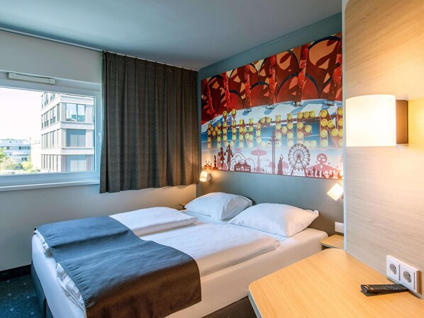 Habitació d'hotel amb un llit, escriptori, finestra amb cortines i paret amb un mural colorit.