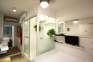 A modern bedroom with a glass shower enclosure, a bed, a bedside table, a TV, and a small plant. - Apple Hotel (Hong Kong Island)