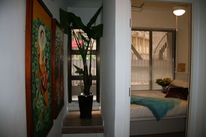 A hallway with a potted plant, a bed with a blue blanket, and a window with blinds. - Apple Hotel (Hong Kong Island)