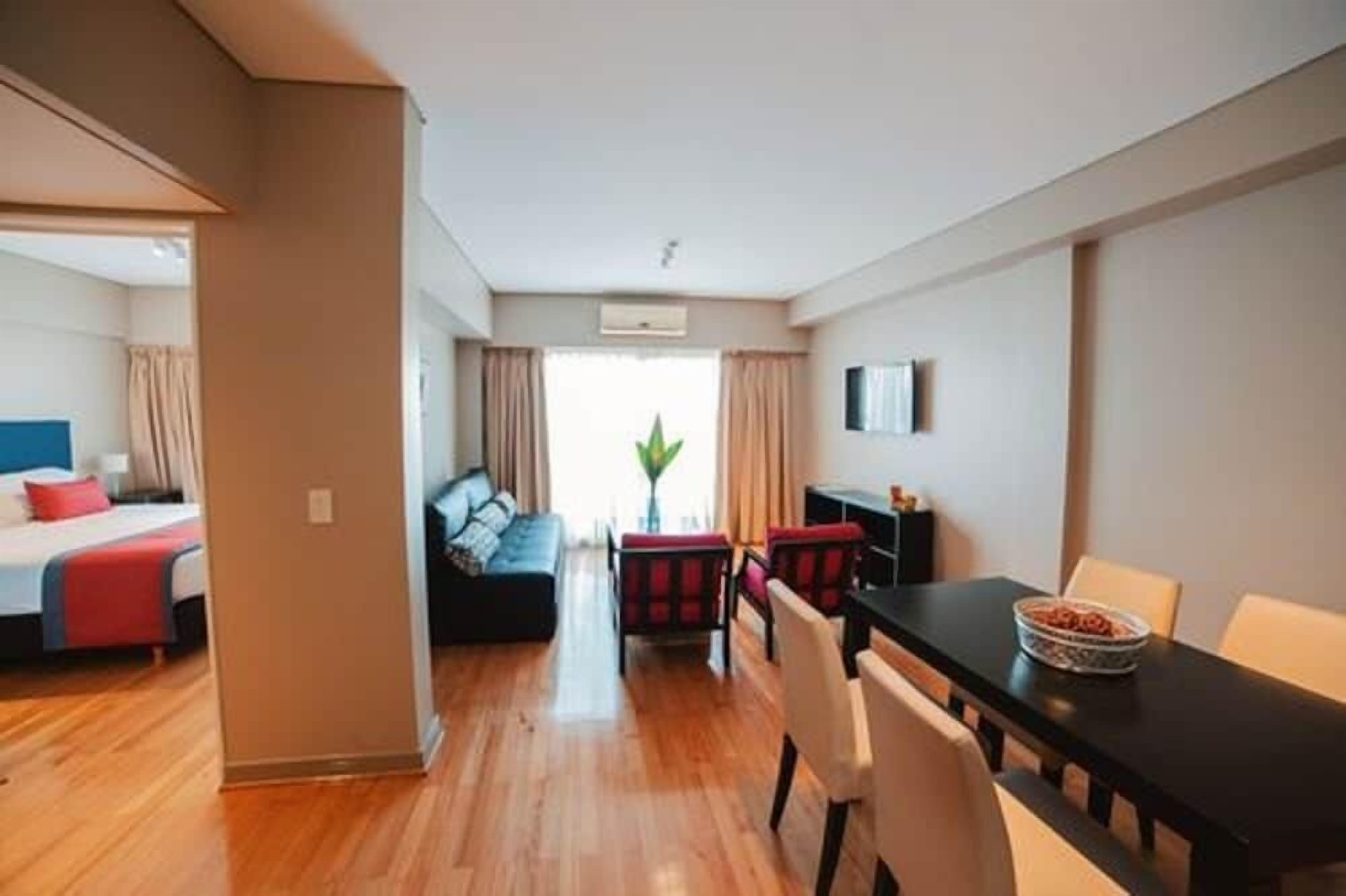 A modern hotel room with a wooden floor, a bed with red bedding, a sofa, a dining table with chairs, and a TV on the wall.